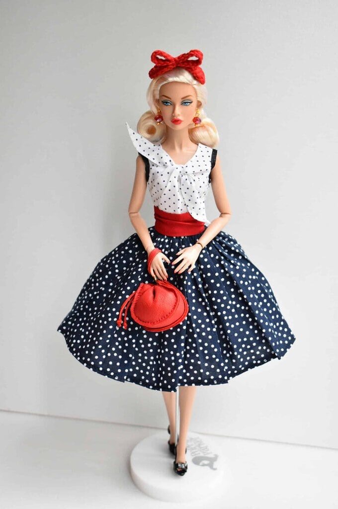 Elegant 1950s-style doll dress featuring a white polka dot top with a bow detail, a fitted red waistband, and a voluminous navy blue polka dot skirt, exuding a timeless pin-up aesthetic.